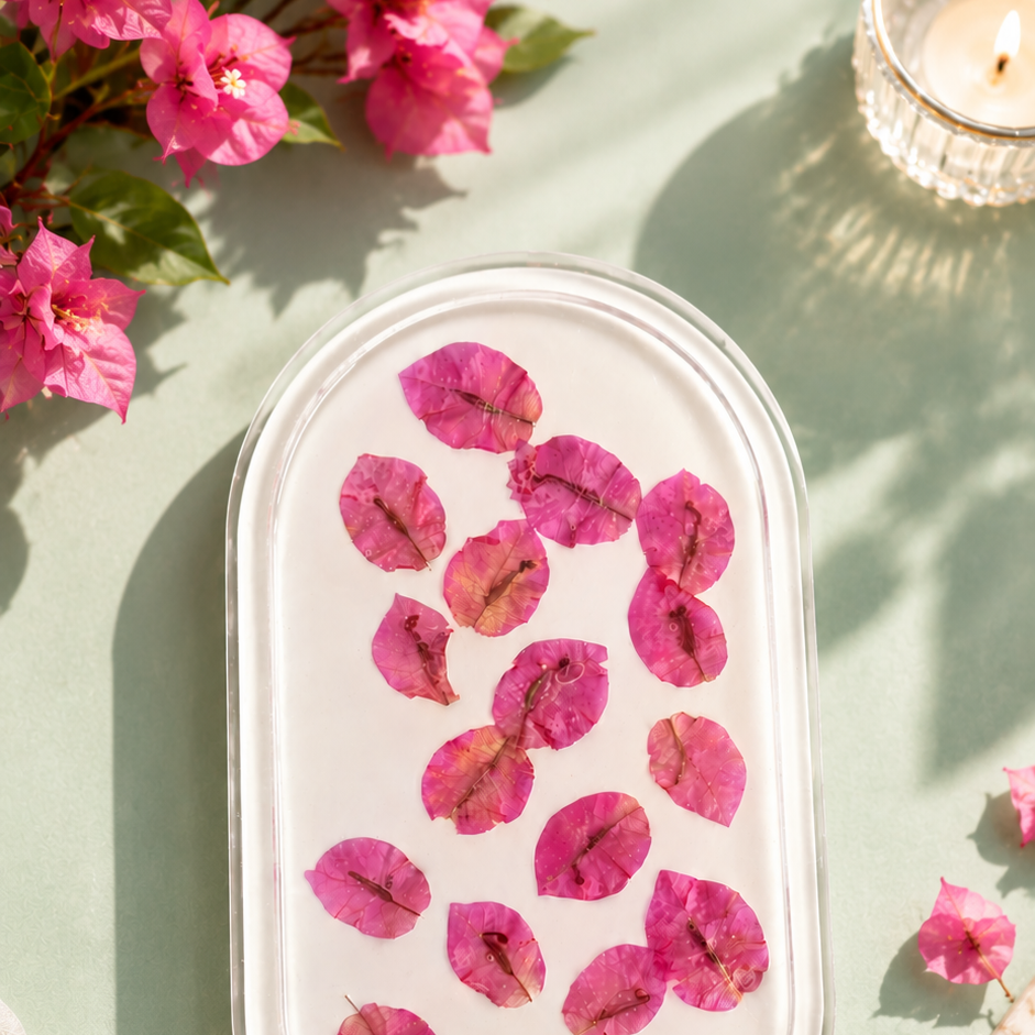 A clear oval glass dish containing scattered pink flower petals, styled on a pale green surface with decorative flowers, a lit candle, and soft fabric in the background.