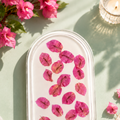 A clear oval glass dish containing scattered pink flower petals, styled on a pale green surface with decorative flowers, a lit candle, and soft fabric in the background.